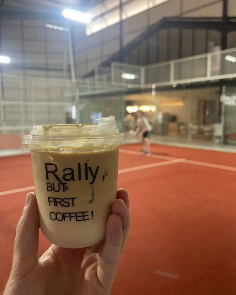 An iced coffee cup printed 'Rally, but first coffee!' held up in front of the court